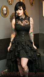 Women's black high low ruffle layer dress in brocade fabric with a sheer lace illusion chest panel and bat shaped collar shown on a model with an additional waist cincher