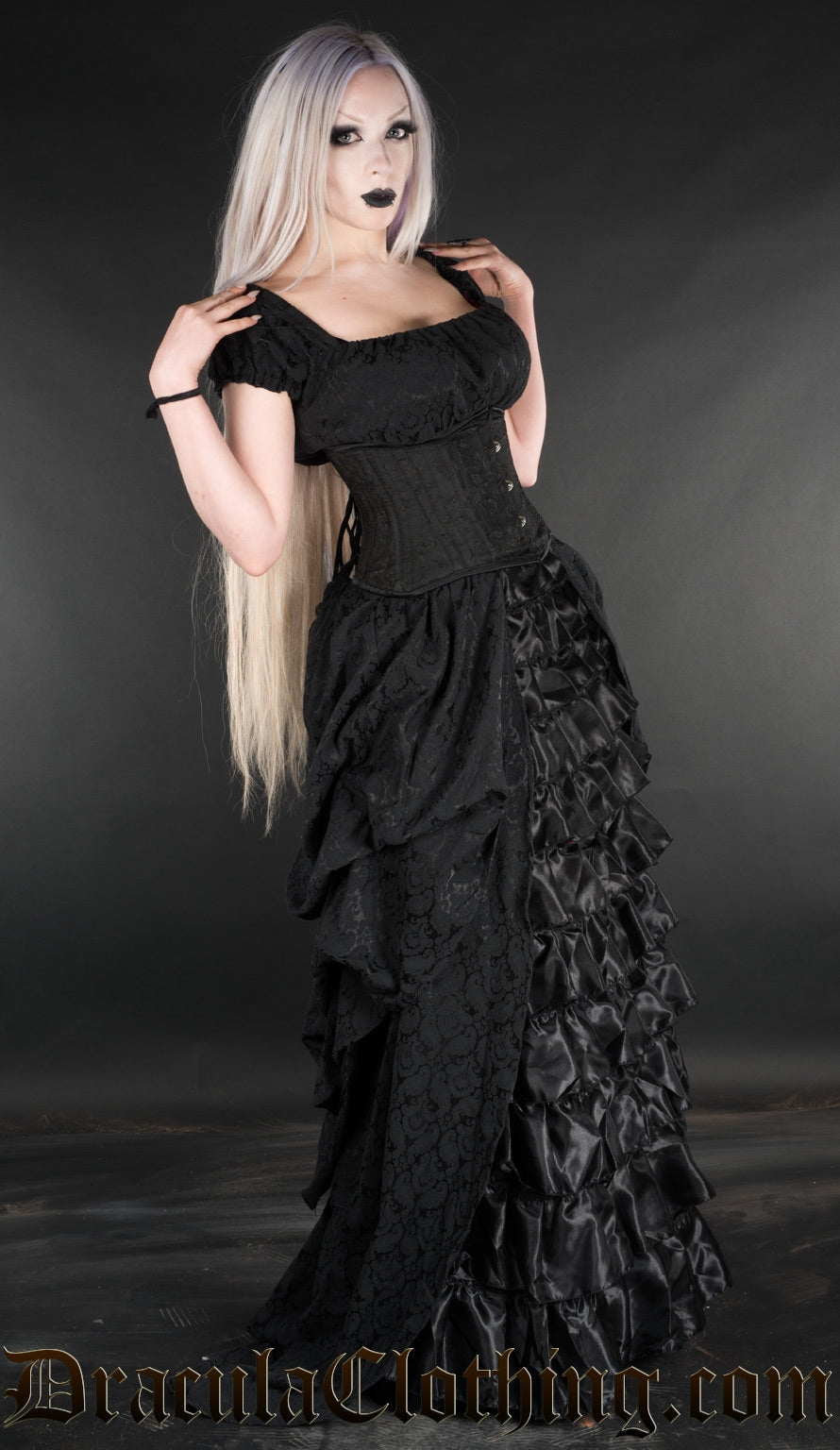 Model wearing short sleeve black full length dress with satin ruffles in the centre of the skirt and black brocade fabric in the back and sides